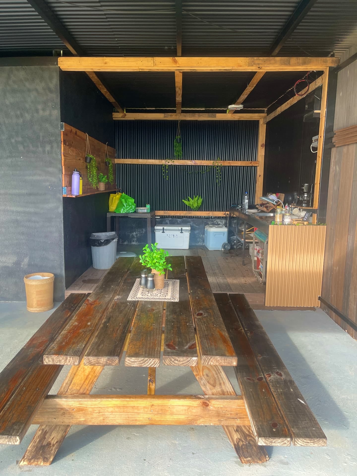 Cocina exterior con mesón, fregadero, cafetera y neveras portátiles, y una mesa de picnic de madera en primer plano