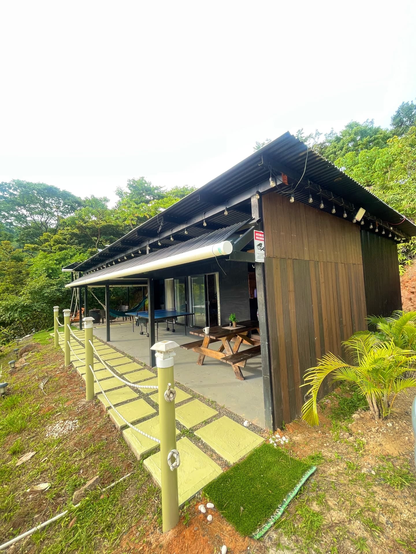 Cabaña de techo oscuro y paredes de madera rodeada de vegetación, con un camino de losas verdes y postes con cuerda
