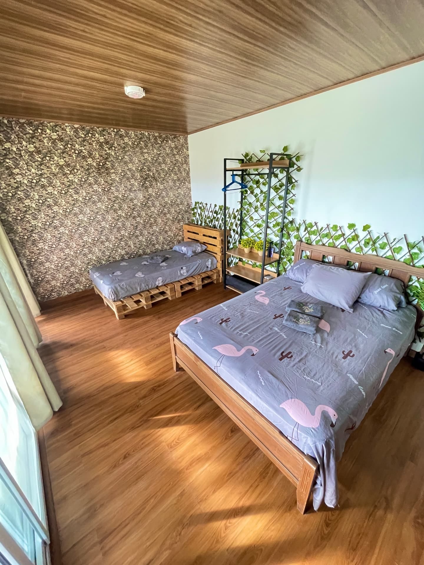 Habitación con techo de madera, cama doble con sábanas de flamencos y una cama sencilla junto a un perchero con plantas