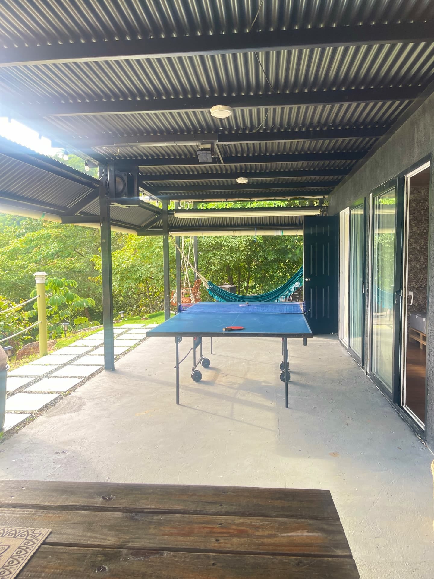 Terraza cubierta con techo de zinc y una mesa de ping-pong azul, con vegetación al fondo y puertas de vidrio a la derecha